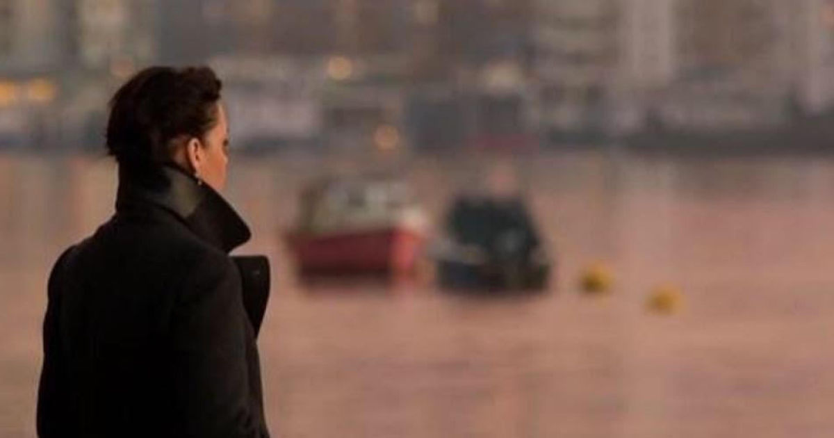 A woman in a black coat looks across a harbour, there are boats in the background